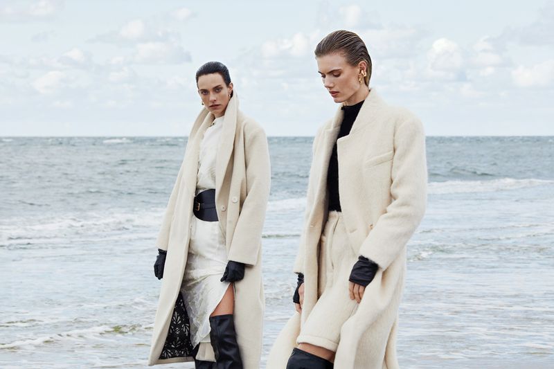 Two women in cream-colored overcoats pose on a beach with a neutral color palette, exuding a high-fashion editorial aesthetic.