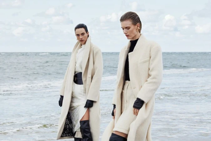 Two women in cream-colored overcoats pose on a beach with a neutral color palette, exuding a high-fashion editorial aesthetic.