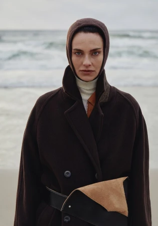 A woman with a neutral expression wears a hooded brown coat with a black belt and light colored sweater on a beach with waves.