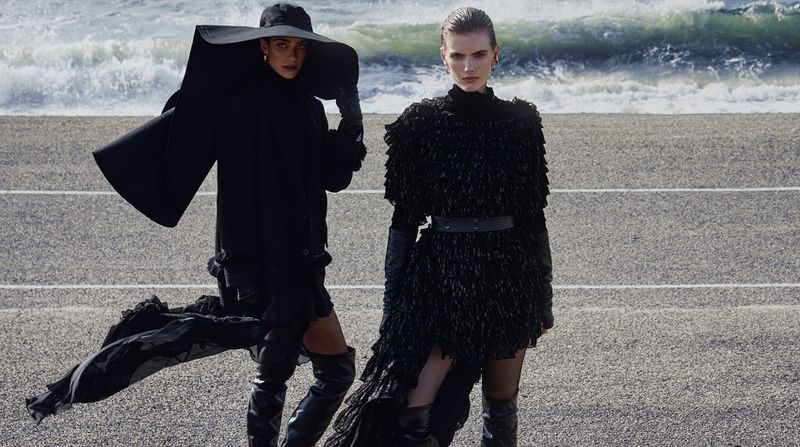 Two models pose in black avant-garde outfits and thigh-high boots on a coastal road with crashing ocean waves in the background.