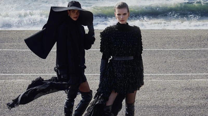 Two models pose in black avant-garde outfits and thigh-high boots on a coastal road with crashing ocean waves in the background.
