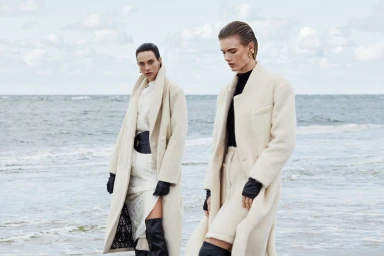 Two fashion models posing in long cream wool coats and black thigh-high boots against an ocean shoreline.