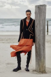 Model posing on a sandy beach beside a wooden post wearing a black blazer with cord trim, orange skirt, and black boots.