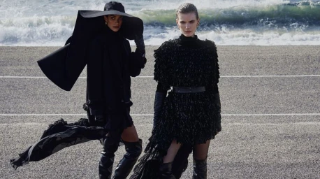 Two models pose in black avant-garde outfits and thigh-high boots on a coastal road with crashing ocean waves in the background.