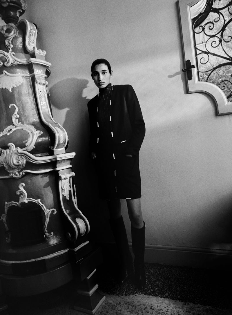 Monochrome fashion shot of a slender adult in a dark coat and knee-high boots, posing next to an ornate plaster column in an indoor setting.