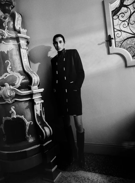 Monochrome fashion shot of a slender adult in a dark coat and knee-high boots, posing next to an ornate plaster column in an indoor setting.