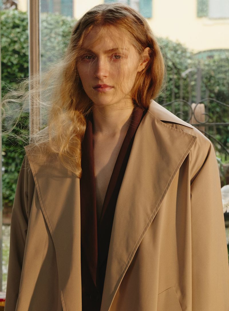 Portrait of a blonde female adult wearing a beige trench coat, posing outdoors with natural light and green foliage visible in the background.