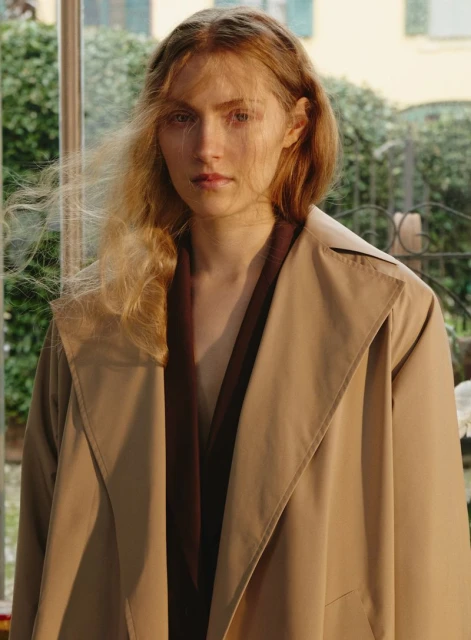 Portrait of a blonde female adult wearing a beige trench coat, posing outdoors with natural light and green foliage visible in the background.