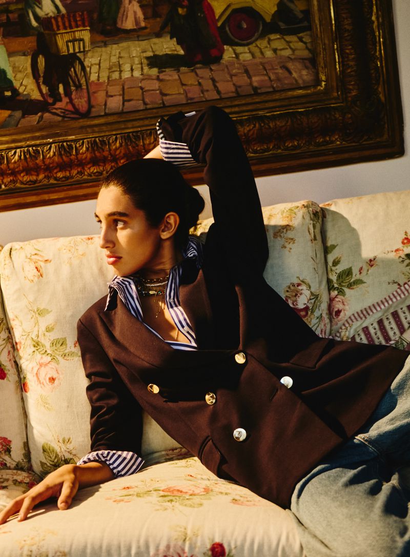 Woman in blazer reclining on floral sofa in front of framed painting. Indoor portrait captures fashion and classic style.