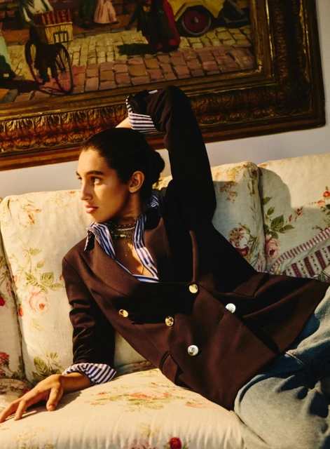 Woman in blazer reclining on floral sofa in front of framed painting. Indoor portrait captures fashion and classic style.