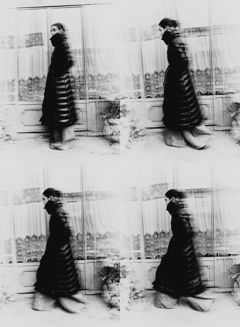Black and white fashion photograph of a woman in a coat, captured in a blurred, time-slice style, standing outdoors against a textured wall.