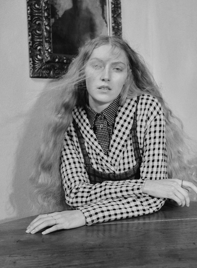 Monochrome portrait of a young woman with long blonde hair, wearing a checkered jacket and patterned shirt, posing indoors.
