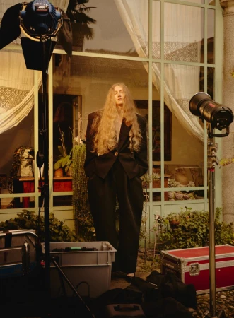 Woman with long blonde hair wearing a black suit stands in front of a window with plants and lighting equipment on either side.