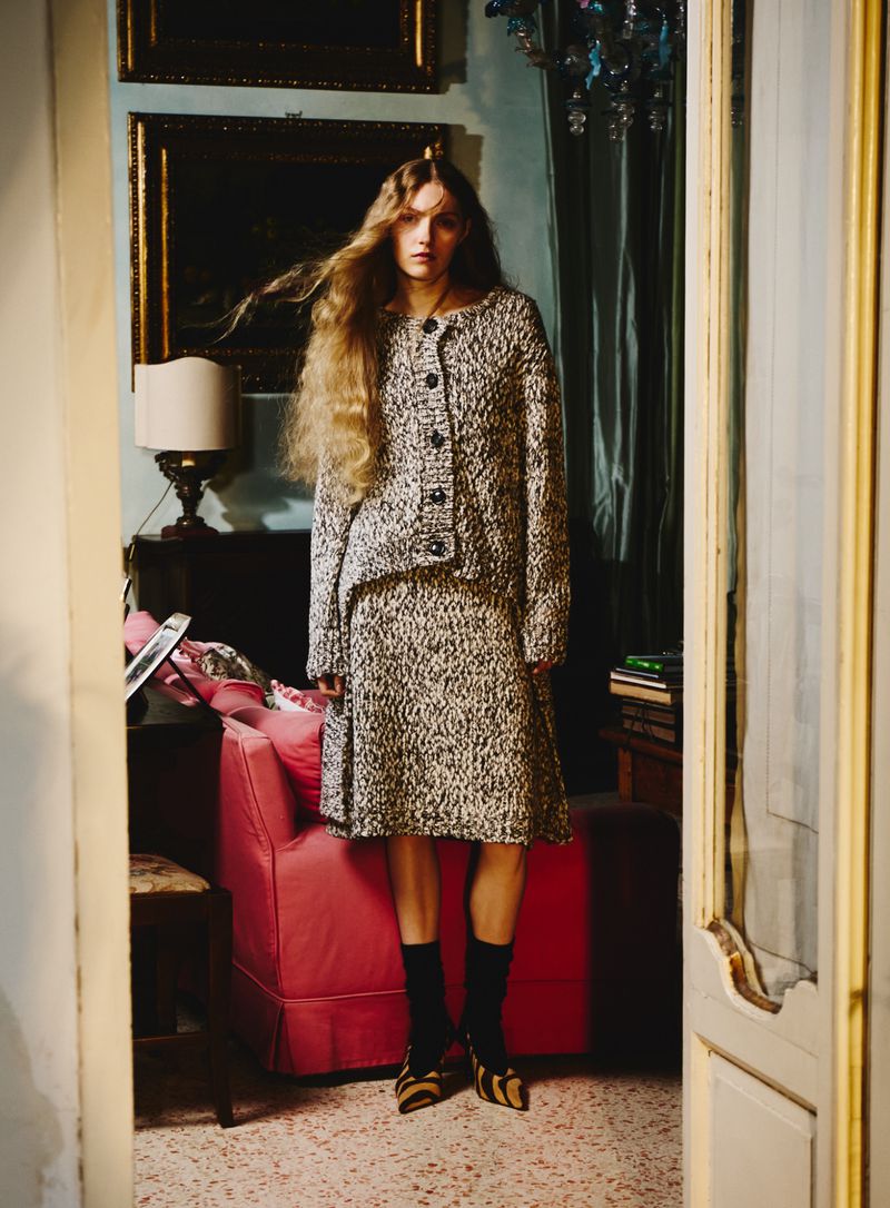 Full shot of a blonde female fashion model wearing a tweed skirt and jacket in an indoor room setting with paintings and furniture.