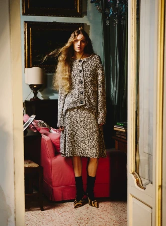 Full shot of a blonde female fashion model wearing a tweed skirt and jacket in an indoor room setting with paintings and furniture.
