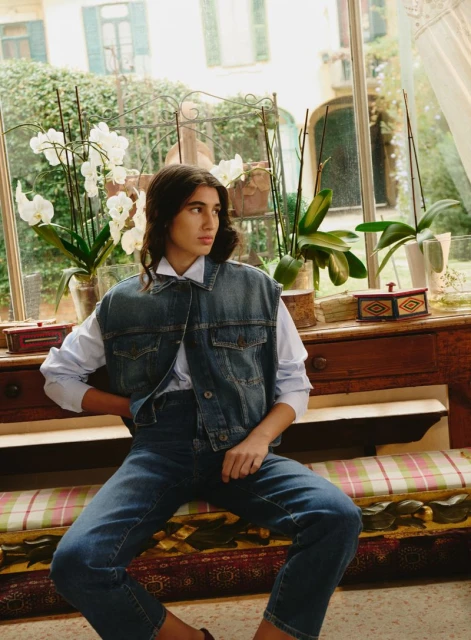 A young woman in a denim vest and jeans sits indoors in front of a window with white orchids, showcasing a casual fashion style.