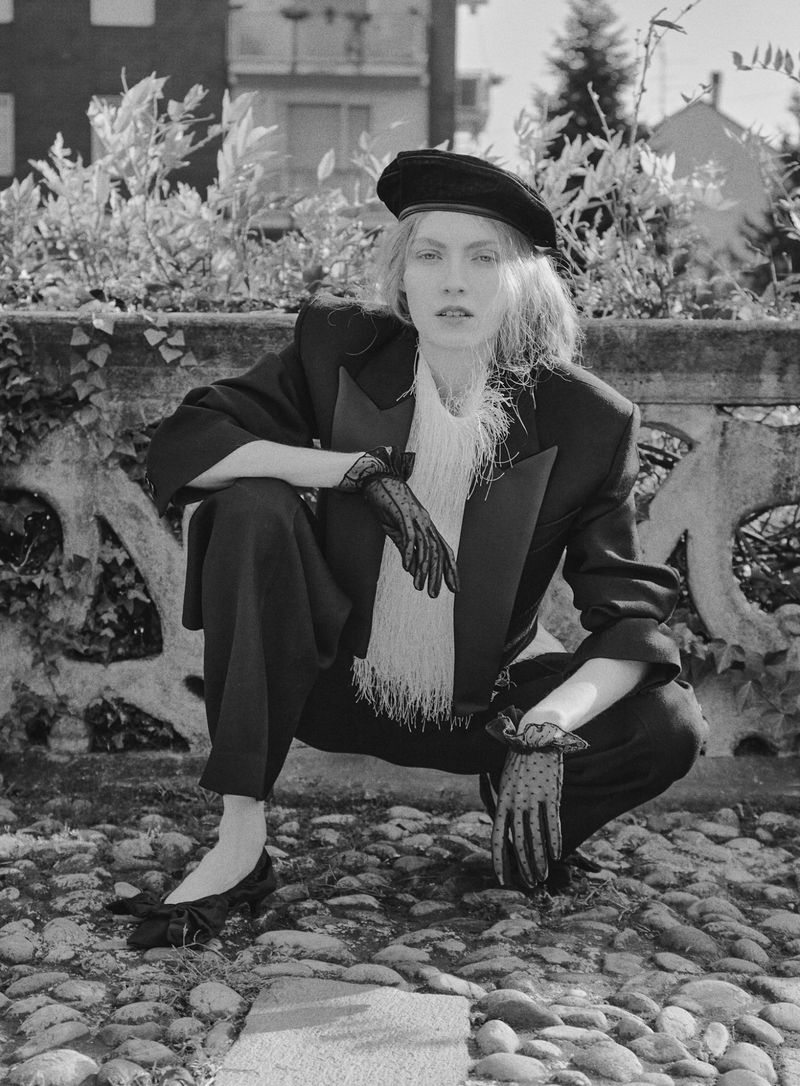 Monochrome portrait of a woman in a black beret and suit, crouching on a cobblestone street, exuding an editorial fashion vibe.