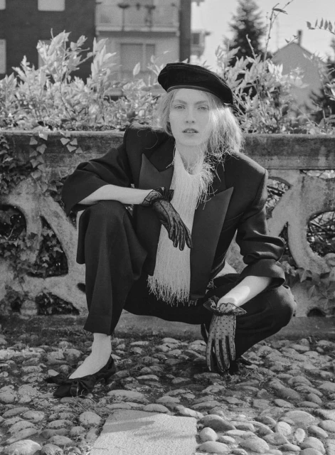 Monochrome portrait of a woman in a black beret and suit, crouching on a cobblestone street, exuding an editorial fashion vibe.