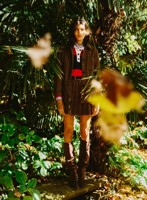 Full-length shot of a woman wearing a brown plaid skirt and jacket, with knee-high boots, standing in a green, leafy outdoor setting.