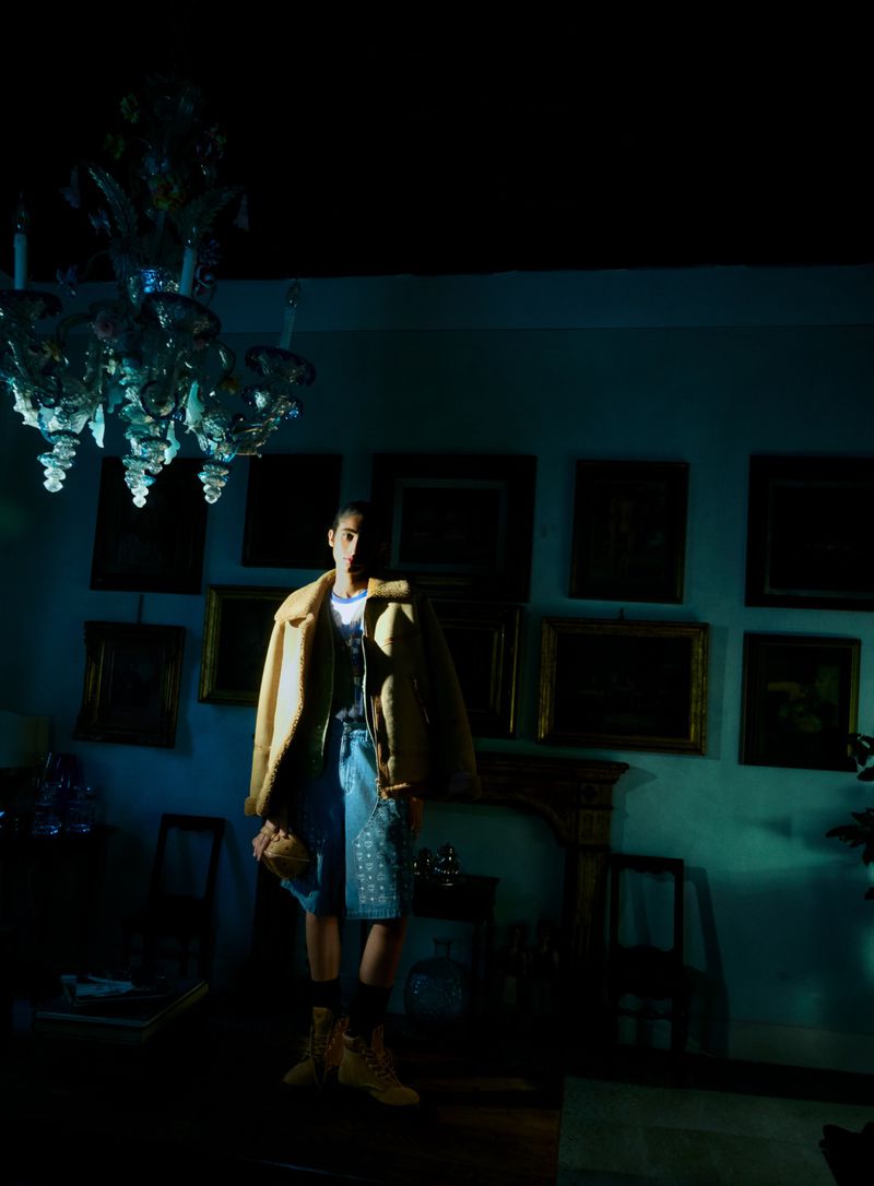 A person in an off-white coat, a top, and shorts poses indoors in a dimly lit room with portraits on the walls and a chandelier.