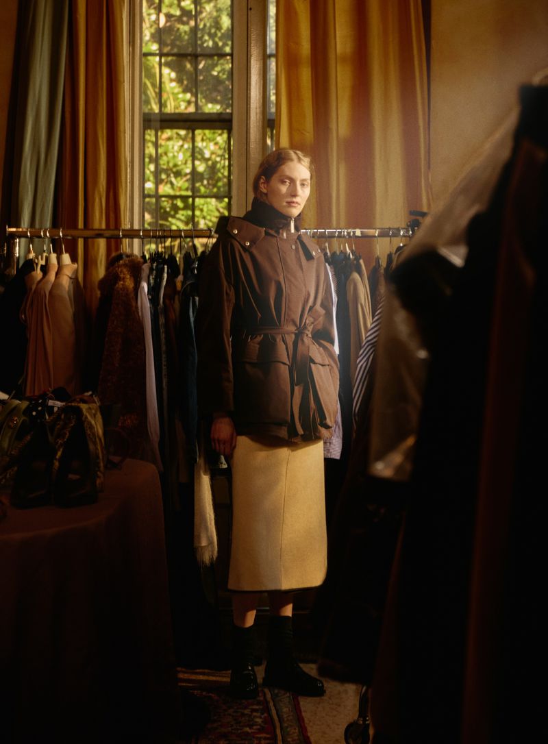 A woman models a brown hooded jacket and beige midi skirt indoors in front of a clothing rack by a window with yellow curtains.