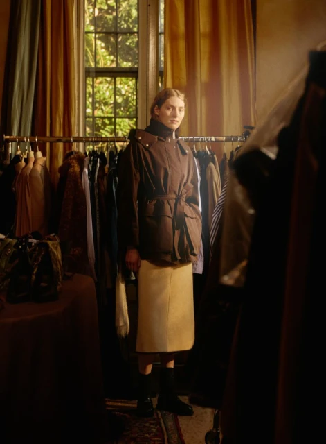A woman models a brown hooded jacket and beige midi skirt indoors in front of a clothing rack by a window with yellow curtains.