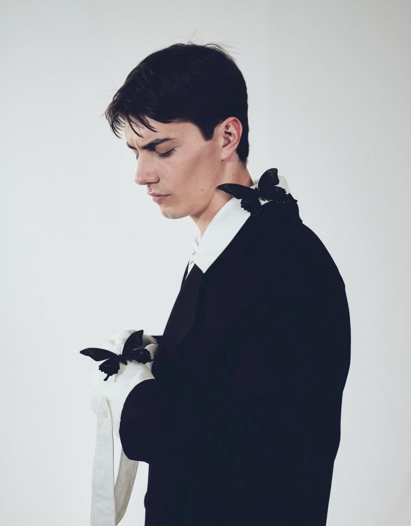 Profile view of a young man in a black suit with a white collared shirt and black butterfly accessories against a white background.