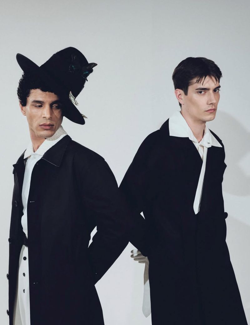 Two adult male fashion models in black coats and white collared shirts pose back-to-back in a photo studio against a white background.