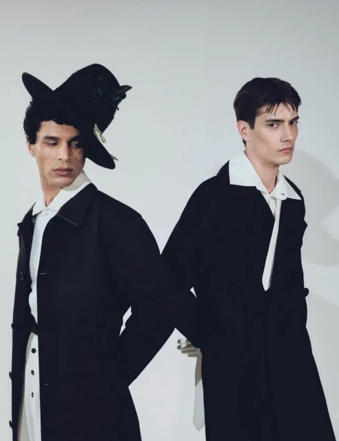 Two adult male fashion models in black coats and white collared shirts pose back-to-back in a photo studio against a white background.