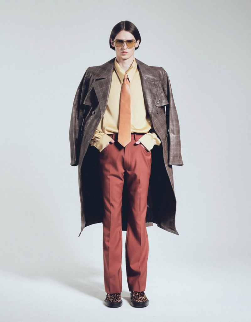 Full-length shot of a male fashion model posing in a yellow collared shirt, brown tie, red-brown pants, and sunglasses, with a brown coat.