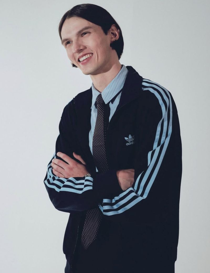 Man in Adidas tracksuit jacket, blue striped shirt, and tie smiles in a studio portrait. Casual style and formal attire mix.
