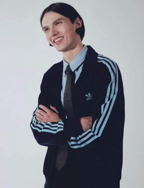 Man in Adidas tracksuit jacket, blue striped shirt, and tie smiles in a studio portrait. Casual style and formal attire mix.