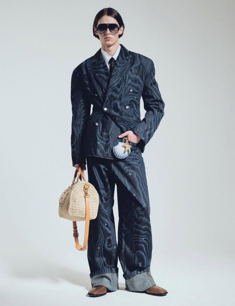 A slender man in a pinstripe double-breasted suit, dress shirt, and tie poses with a straw bag and seashell coin purse on a light backdrop.