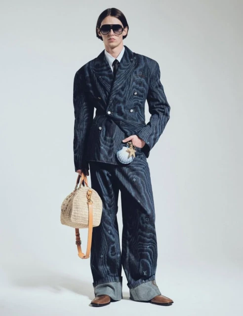 A slender man in a pinstripe double-breasted suit, dress shirt, and tie poses with a straw bag and seashell coin purse on a light backdrop.