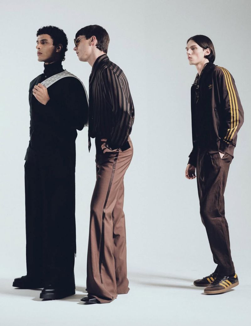 Three adult male fashion models in a studio setting, showcasing modern clothing with black, brown, and beige color palettes, standing in a line.