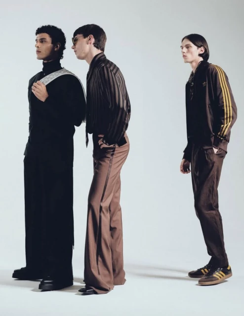 Three adult male fashion models in a studio setting, showcasing modern clothing with black, brown, and beige color palettes, standing in a line.