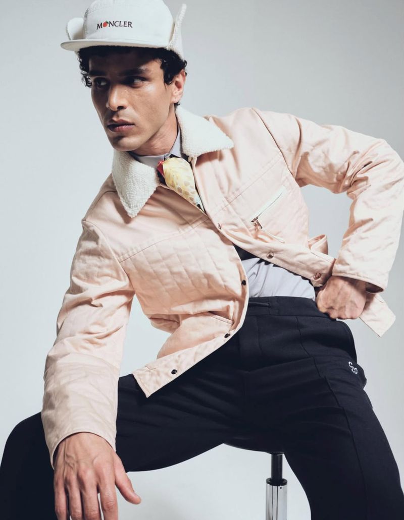 A light-skinned male fashion model poses while seated, wearing a light pink jacket, black pants, and a Moncler baseball cap.