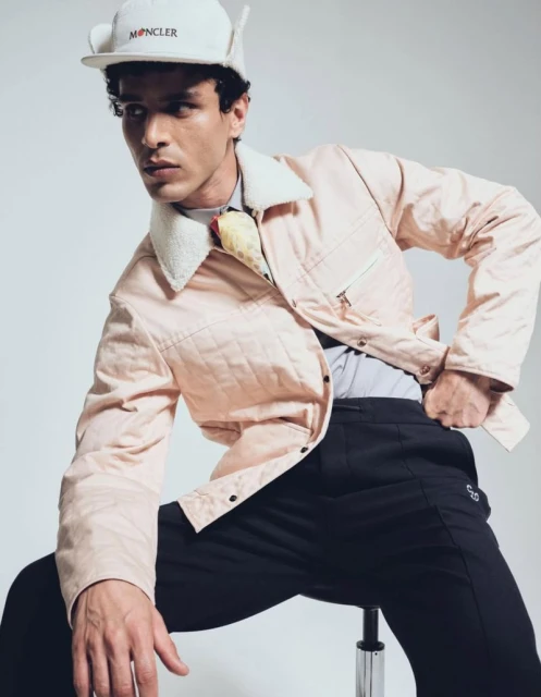 A light-skinned male fashion model poses while seated, wearing a light pink jacket, black pants, and a Moncler baseball cap.