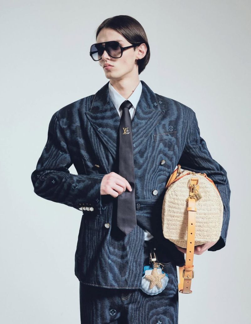 Male fashion model in a blue patterned suit, tie, and sunglasses, holding a tan woven bag with a seashell keychain against a light background.