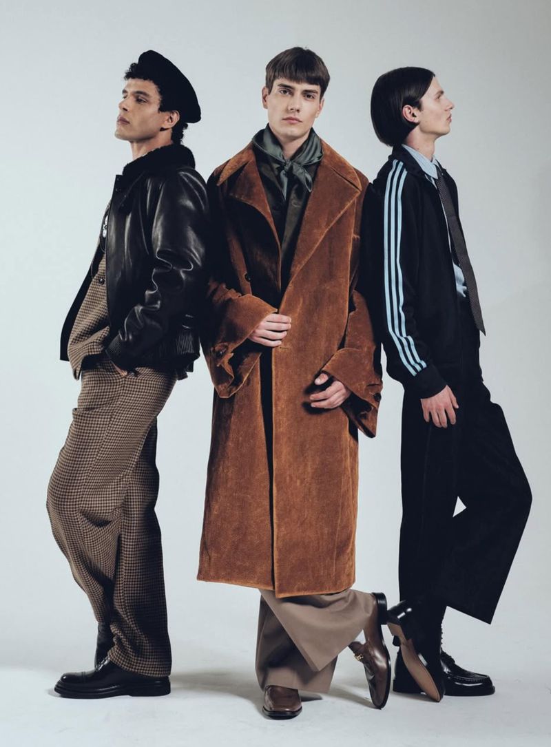 Three male fashion models strike a pose indoors against a clean white background, showcasing an array of jackets, pants, and footwear.