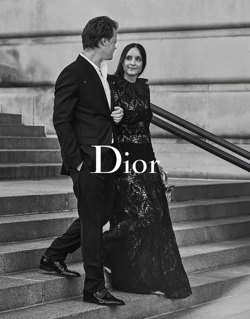 Monochrome image capturing a couple on steps, the woman in a lace dress, the man in a suit, Dior logo overlay, fashion-forward.