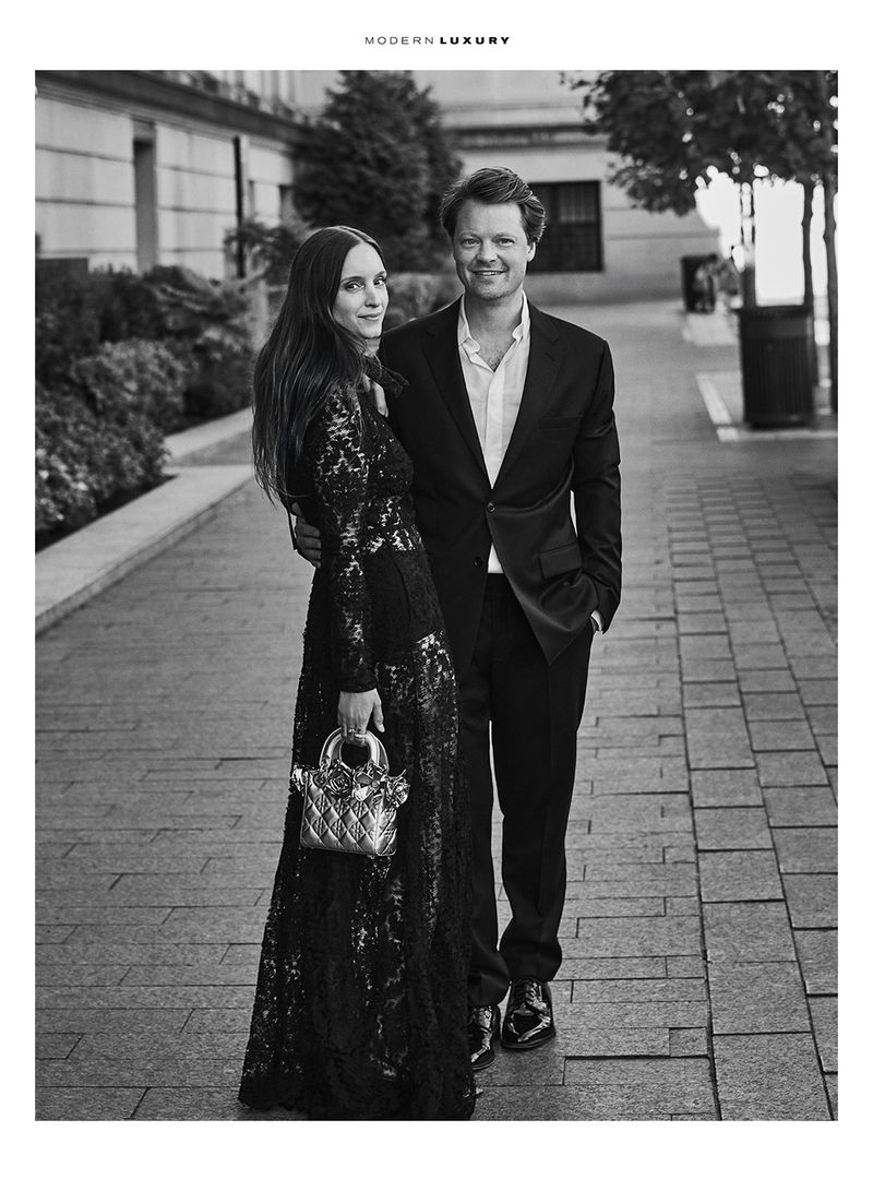 Monochrome portrait of a stylish couple outdoors with building backdrop. The woman has a sequined gown and purse. The man wears a suit.