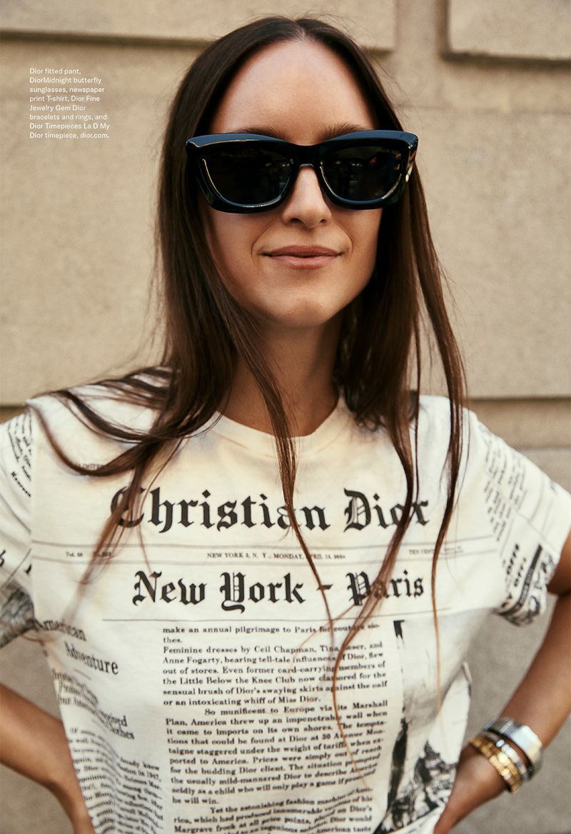 A woman wears sunglasses and a Christian Dior newspaper-print t-shirt while posing outdoors against an unidentifiable beige building.
