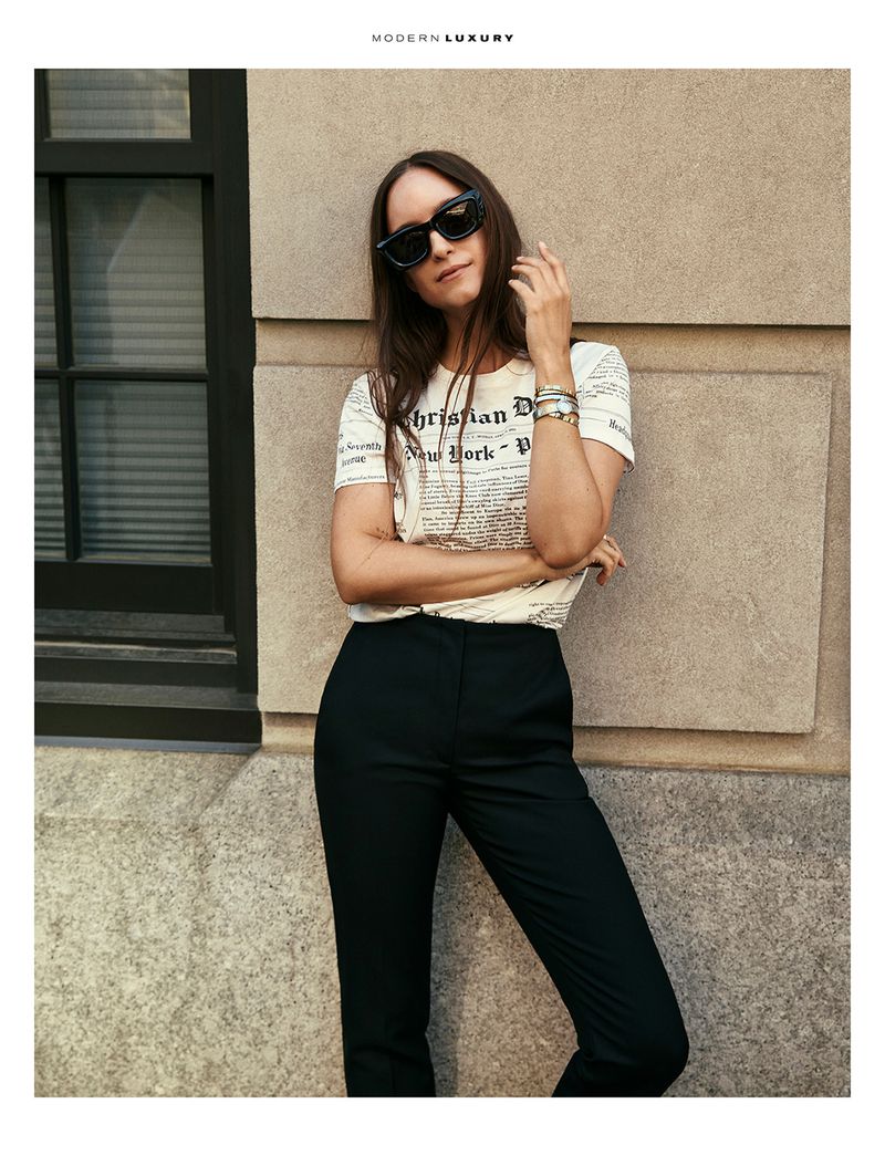 A woman in sunglasses stands in front of a stone wall wearing a white graphic t-shirt and black pants in an urban setting.