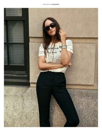 A woman in sunglasses stands in front of a stone wall wearing a white graphic t-shirt and black pants in an urban setting.