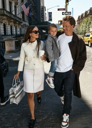 Family strolls through a sunlit urban street. A woman in a cream suit, a man in jeans, and a child are captured in a candid moment.