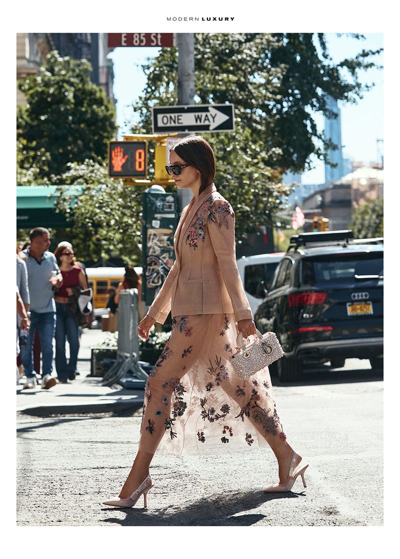 Fashionable woman in floral suit crossing a New York City street. She's holding a handbag and wearing high heels, with urban background.