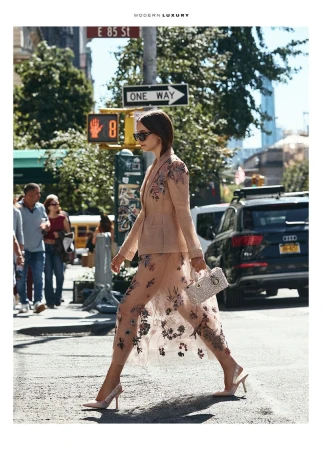 Fashionable woman in floral suit crossing a New York City street. She's holding a handbag and wearing high heels, with urban background.