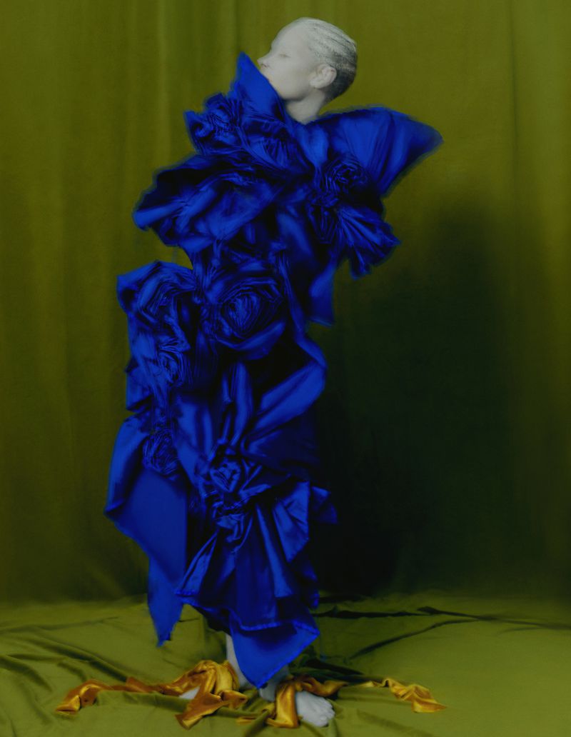 Abstract fashion shot of a model wearing a ruffled, royal blue garment against a muted green backdrop with gold fabric accents.