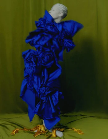 Abstract fashion shot of a model wearing a ruffled, royal blue garment against a muted green backdrop with gold fabric accents.